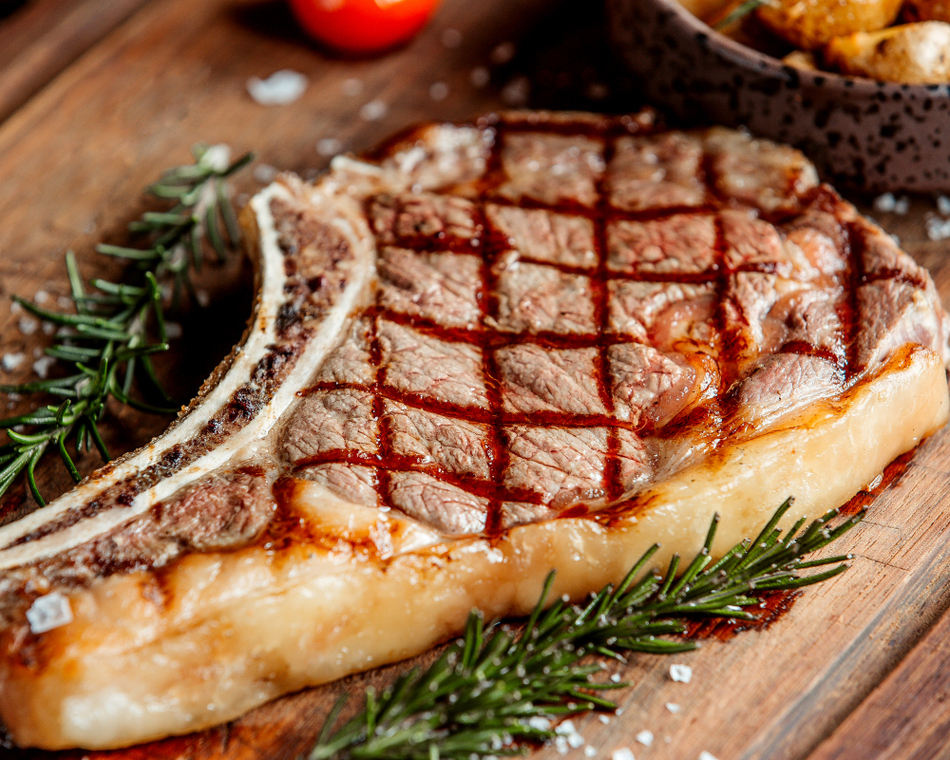 Bone-in ribeye steak with crosshatch grill marks on a wooden board, garnished with rosemary sprigs.