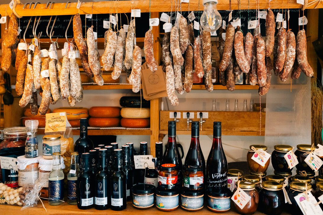 Rustic market stall with numerous hanging cured sausages above a counter of cheese, jars, and bottles like wine and preserves.
