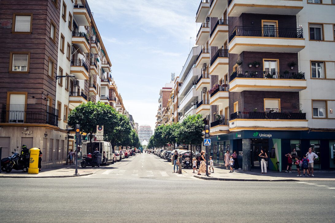 781457 Fincas Legio - León Digital City street with tall residential buildings on both sides, pedestrians crossing a crosswalk and cars parked along the curb under a blue sky.
