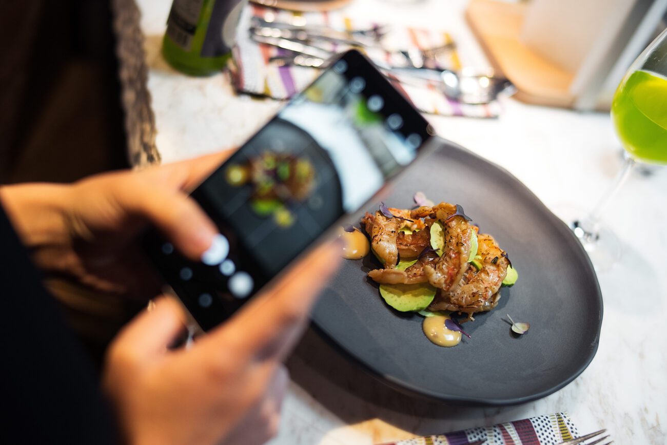 Young woman is holding mobile phone and making photo of her meal - León Digital Person holds a smartphone and photographs a plated gourmet dish on a dark oval plate.
